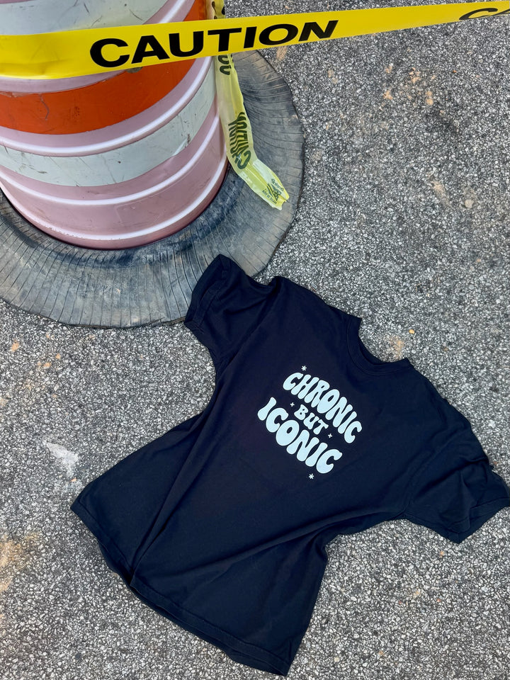 Black t-shirt with 'Chronic but Iconic' text on a concrete surface with caution tape and road barrier in the background.