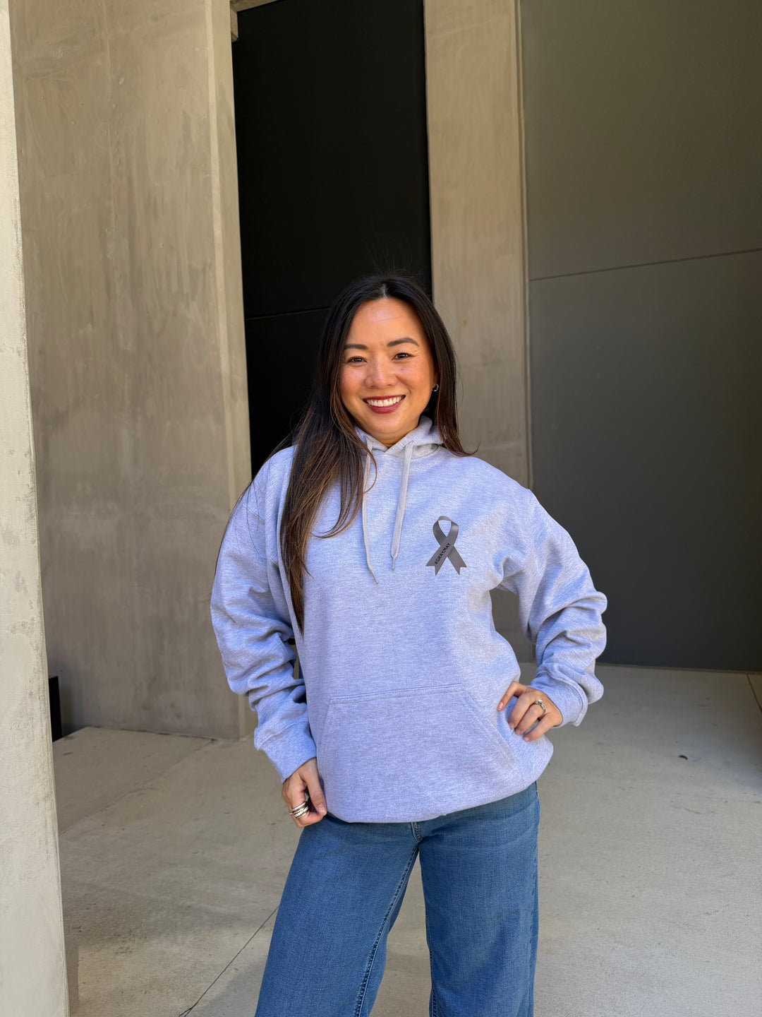 Person wearing a gray sweatshirt with a ribbon design on it, standing in front of a wall. Ribbon has #graymay on it.