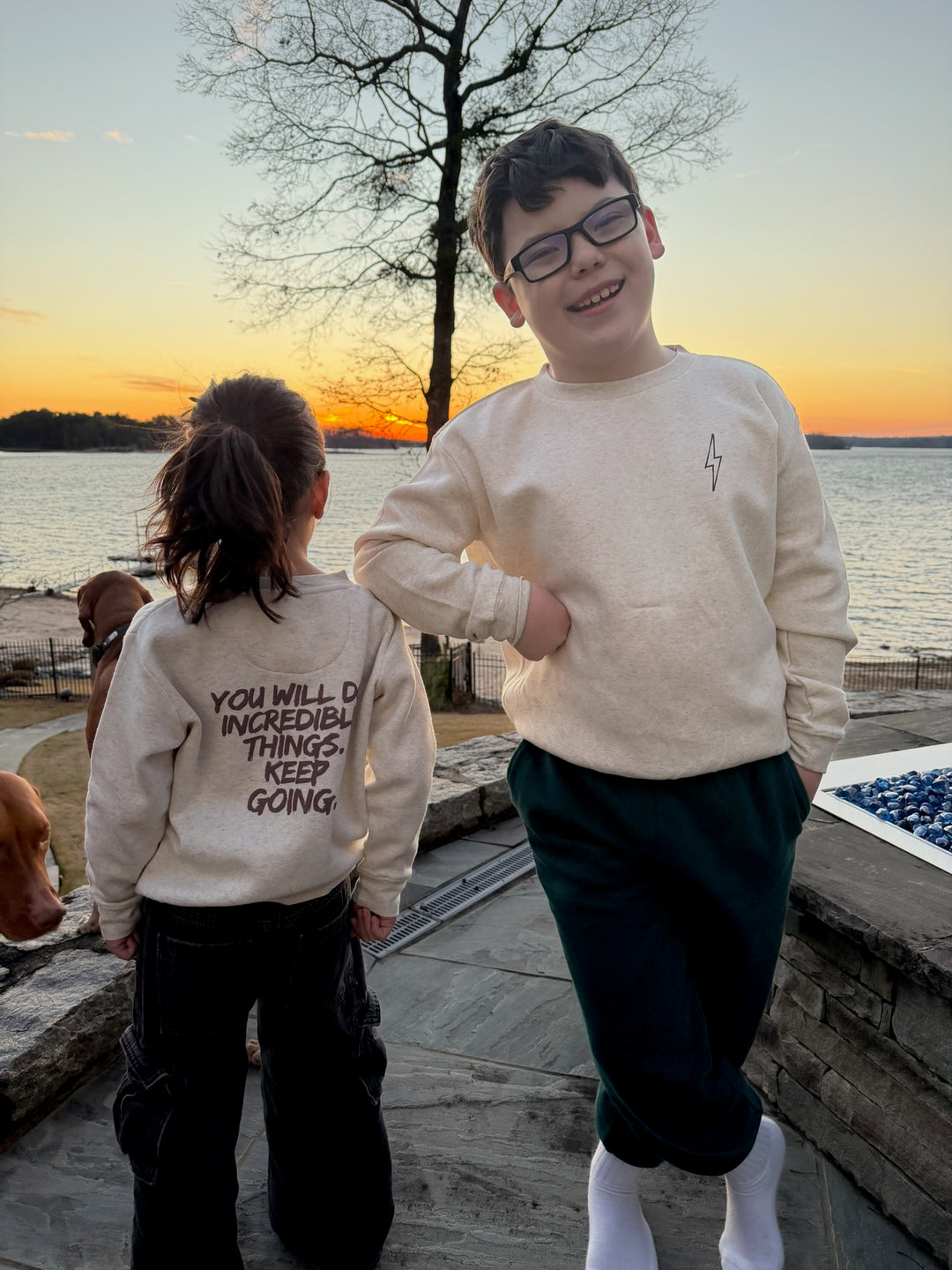 Two children standing by a lake at sunset, wearing The Incredible Crewneck Youth Sweatshirt - Kids Natural Heather Shirt with Motivational "YOU WILL DO INCREDIBLE THINGS. KEEP GOING." Motto