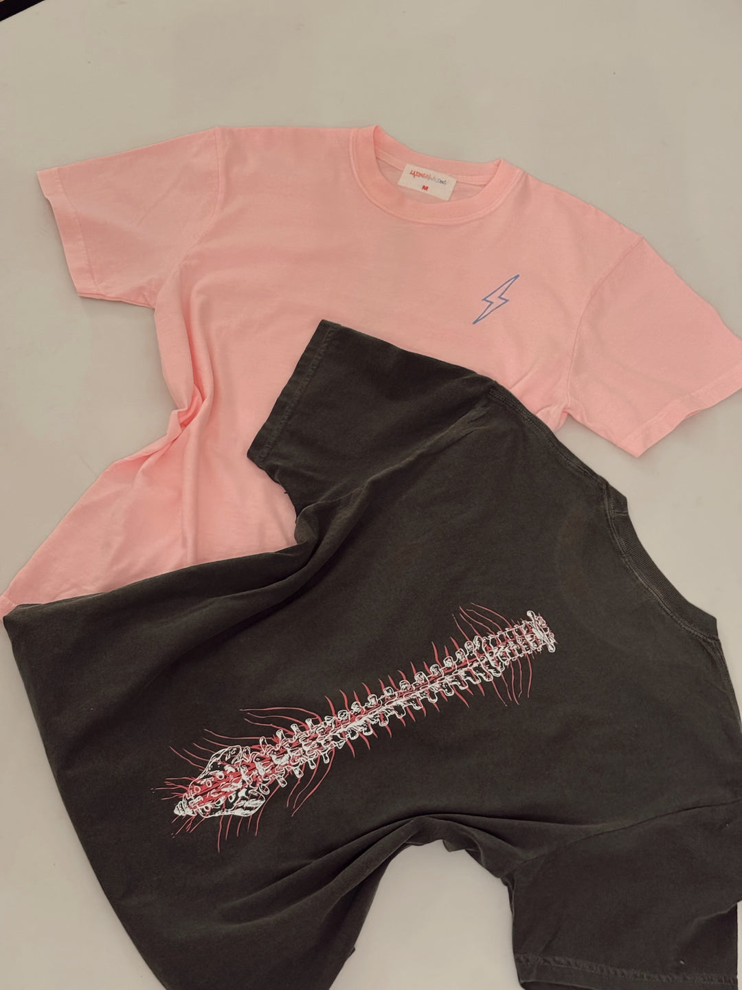Two t-shirts, one pink with a blue lightning bolt, and one black with a detailed red and white spine design on the back.