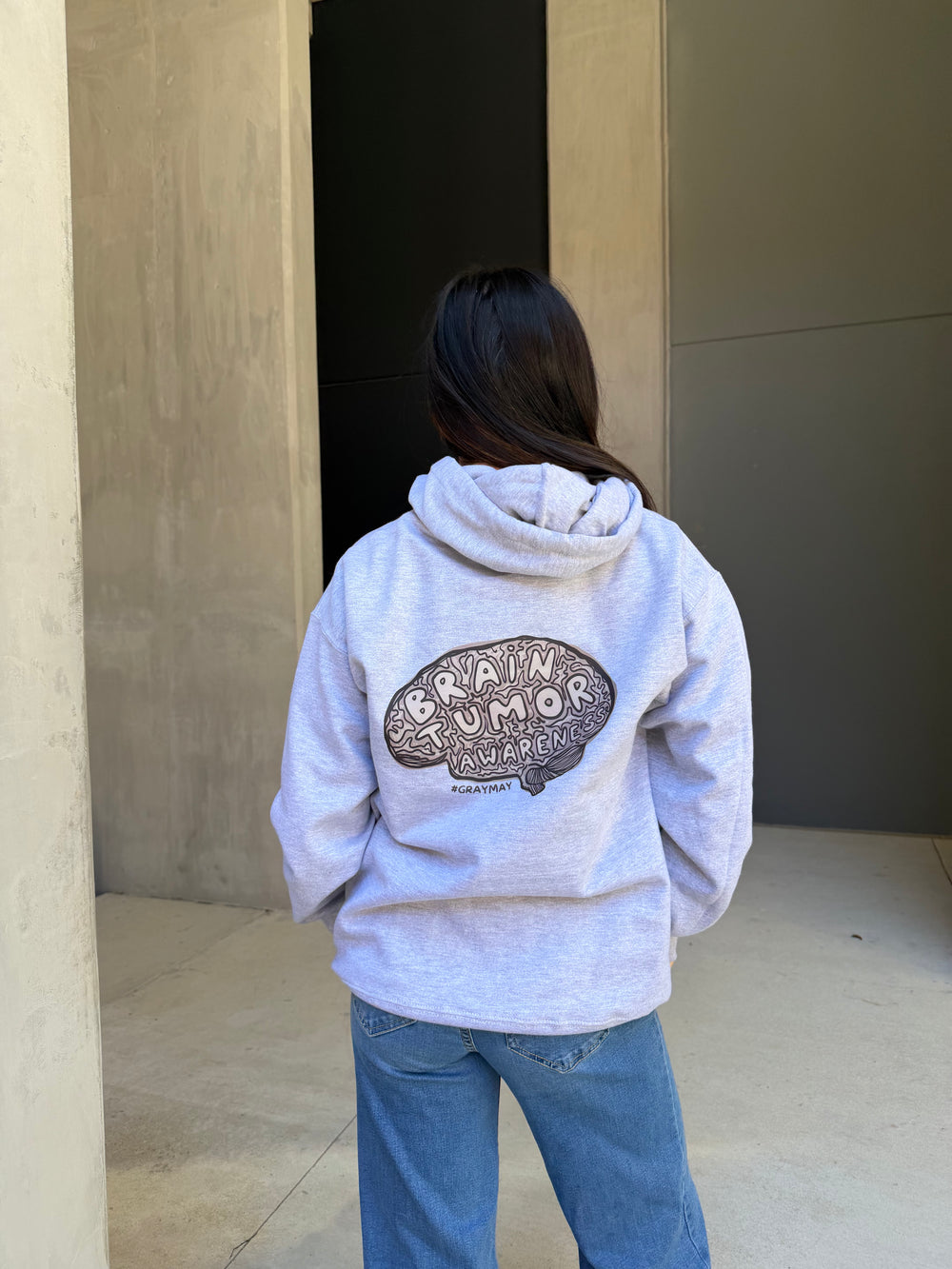 Person wearing a gray hoodie with a brain tumor awareness logo, standing outside a building.