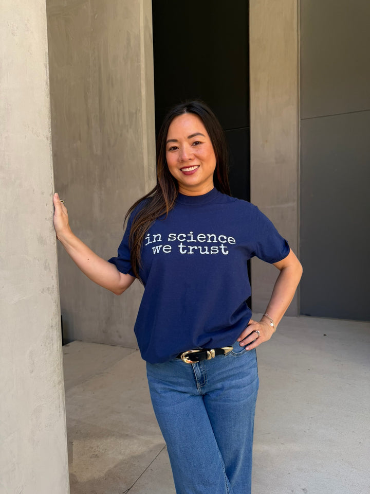 Person wearing a blue t-shirt with 'In science we trust' text, standing against a plain wall.