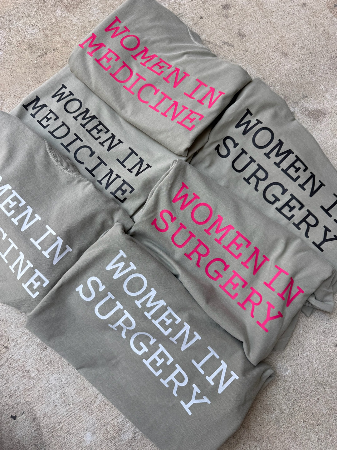 Gray shirts with 'Women in Medicine' and 'Women in Surgery' text on a concrete surface