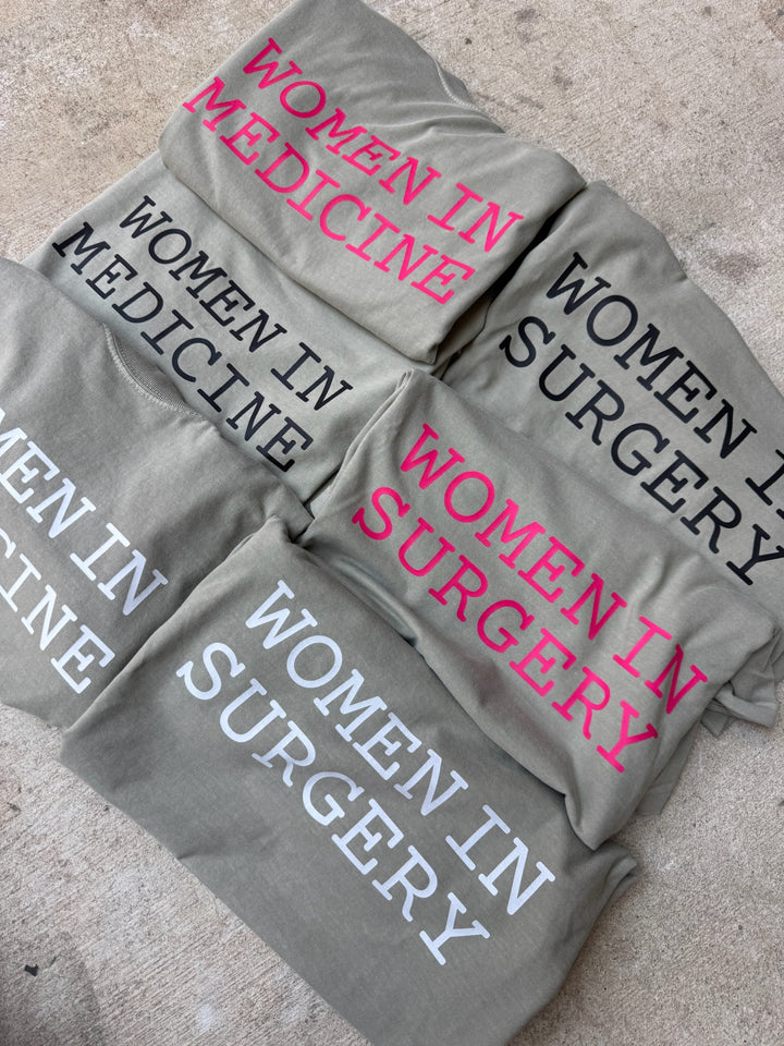 Gray shirts with 'Women in Medicine' and 'Women in Surgery' text on a concrete surface