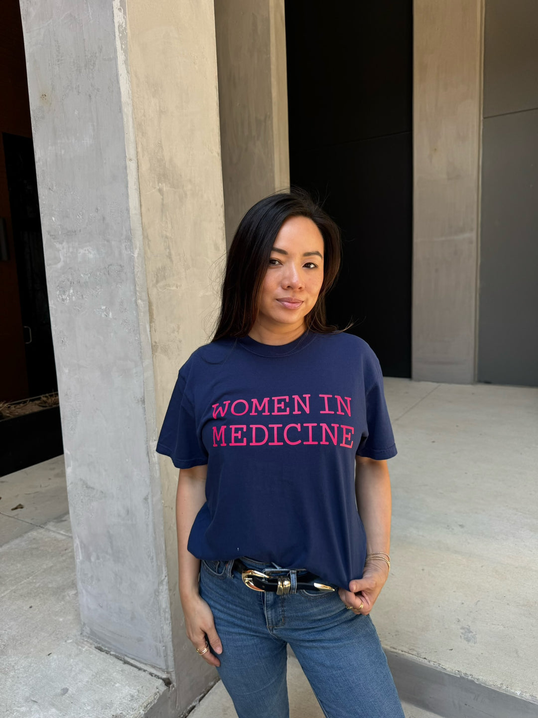 Person wearing a navy blue t-shirt with 'Women in Medicine' in pink text, standing against a concrete wall.