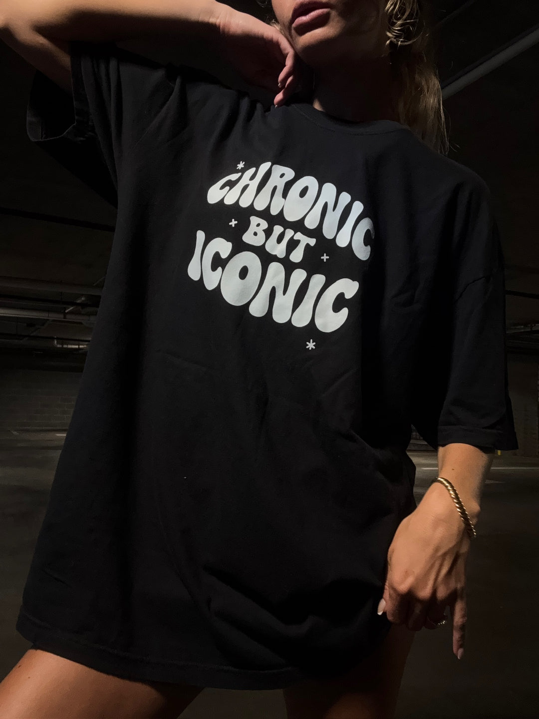 Person wearing a black t-shirt with 'Chronic But Iconic' text in a dark setting