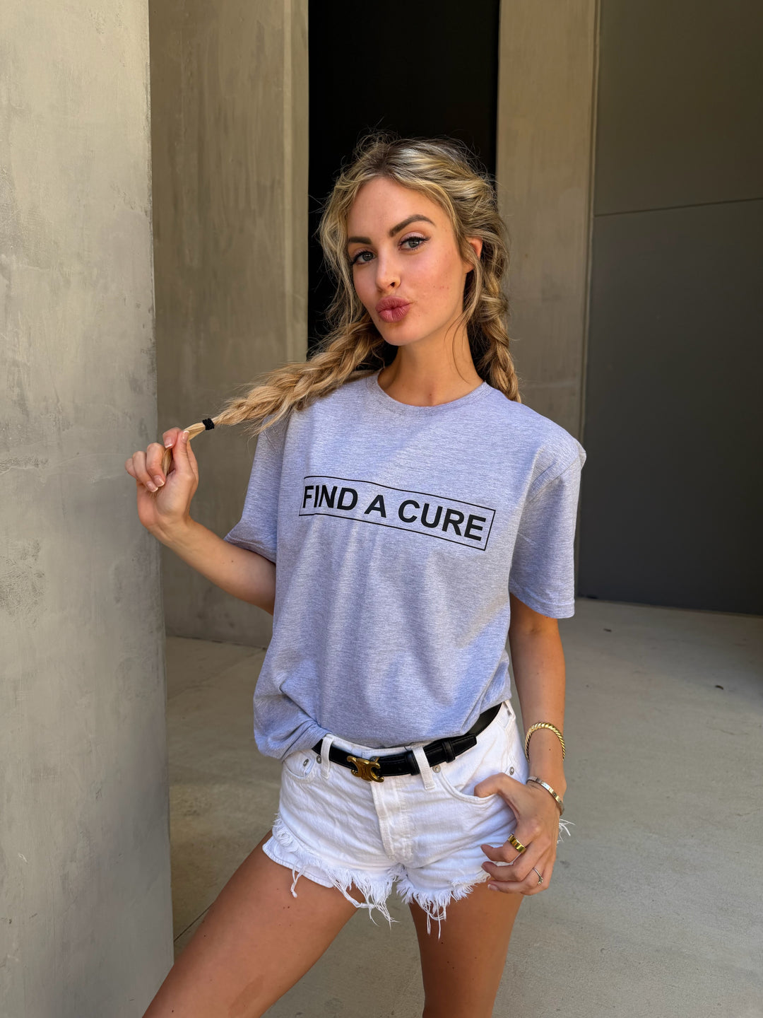 Woman wearing a gray t-shirt with 'Find a Cure' text, standing against a concrete wall.