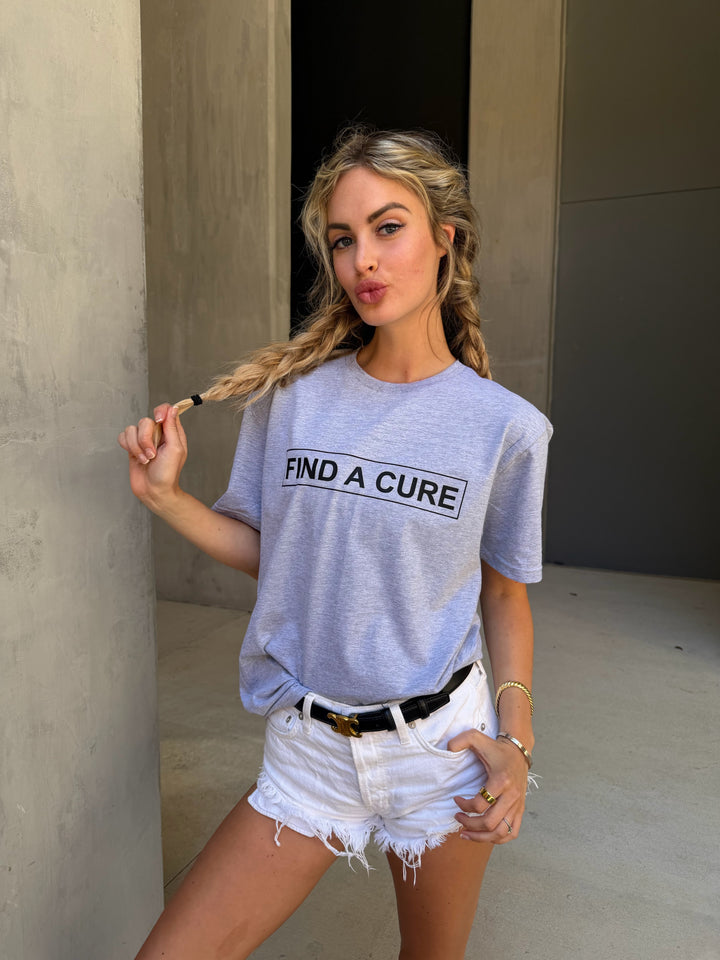 Woman wearing a gray t-shirt with 'Find a Cure' text, standing against a concrete wall.