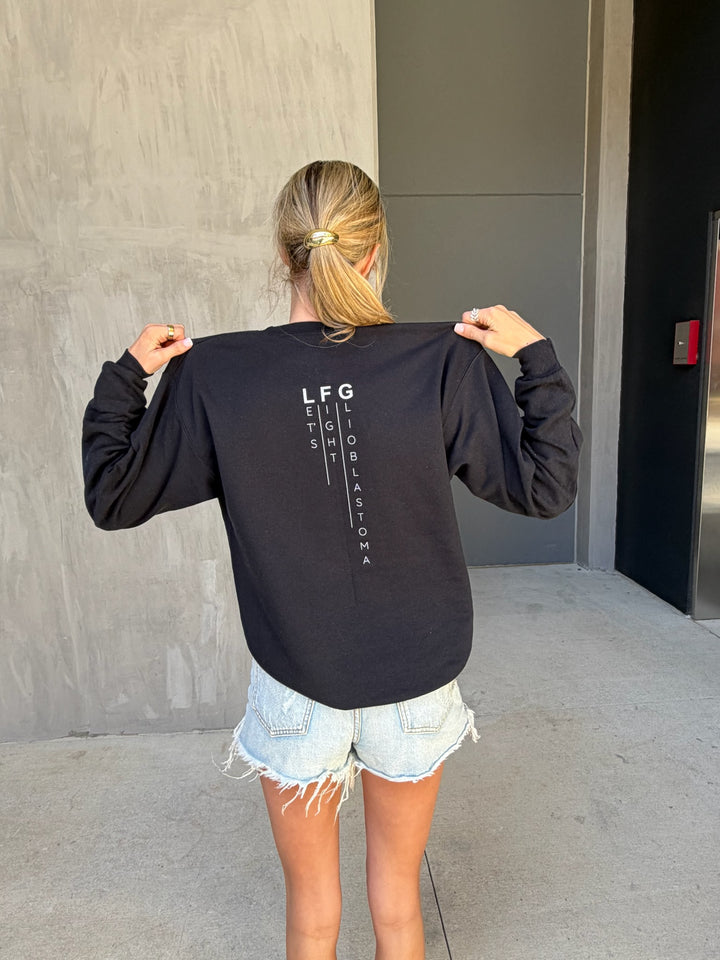 Person wearing a black sweatshirt with Let's Fight Glioblastoma text on the back, standing against a gray wall.