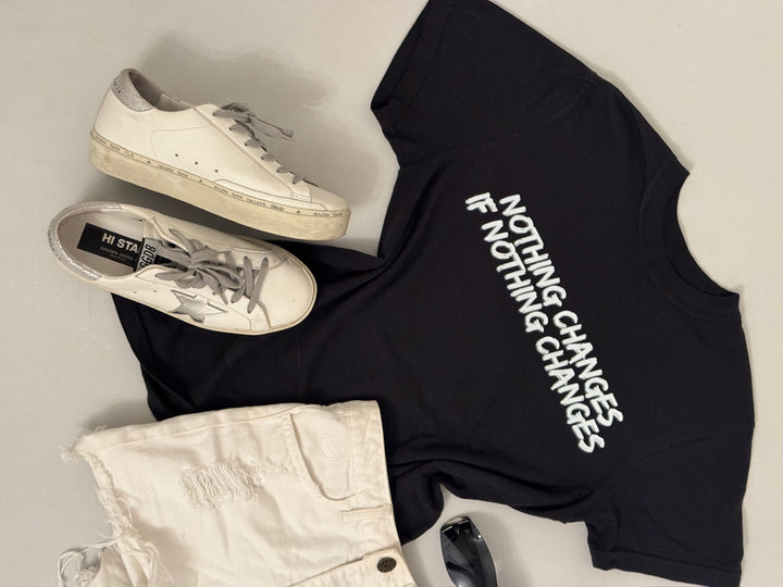 Black Nothing Changes If Nothing Changes shirt laid flat with white sneakers, and light-colored shorts on a neutral background