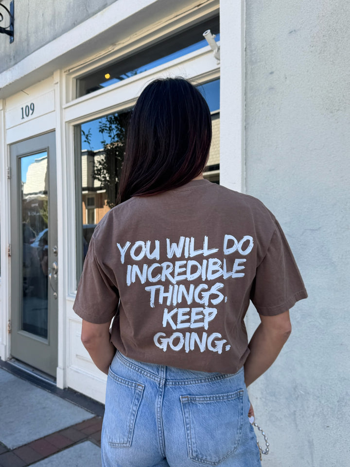 Person wearing The Incredible Tee Shirt - Brown Shirt with Motivational "YOU WILL DO INCREDIBLE THINGS. KEEP GOING." Motto, standing outside a building.