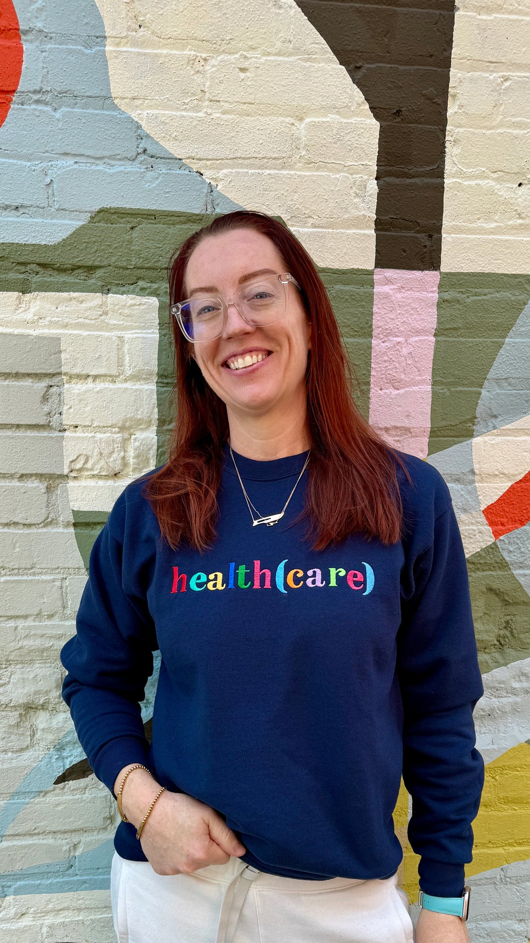Ladyspinedoc wearing a navy blue sweatshirt with 'health(care)' printed on it, standing in front of a colorful mural.