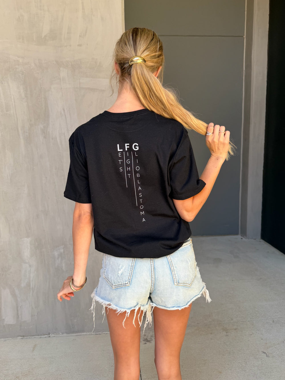 Person wearing a black t-shirt with LFG (Let's Fight Glioblastoma) text on the back, standing against a plain wall.