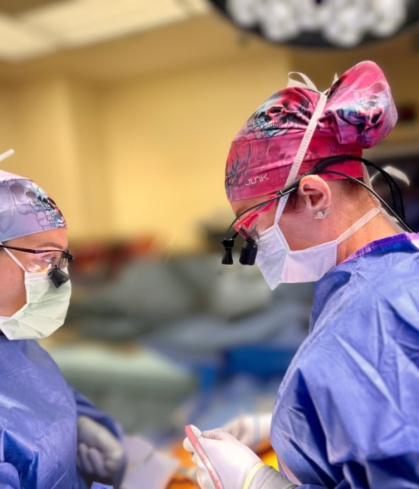 Two surgeons in an operating room, wearing Ladyspinedoc scrub caps, and standard masks and scrubs.