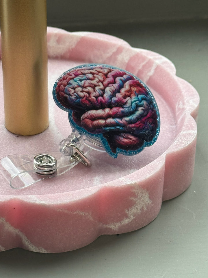 Colorful anatomical brain badge reel with pink and blue design on glittery blue background and retractable clip displayed in a pink scalloped tray.