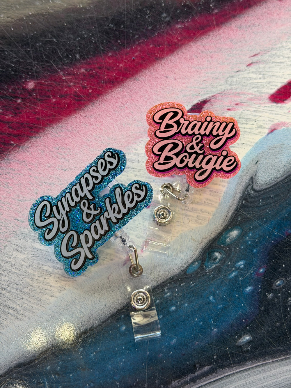 Two decorative badge reels on a textured surface with 'Synapses & Sparkles' and 'Brainy & Bougie' text.