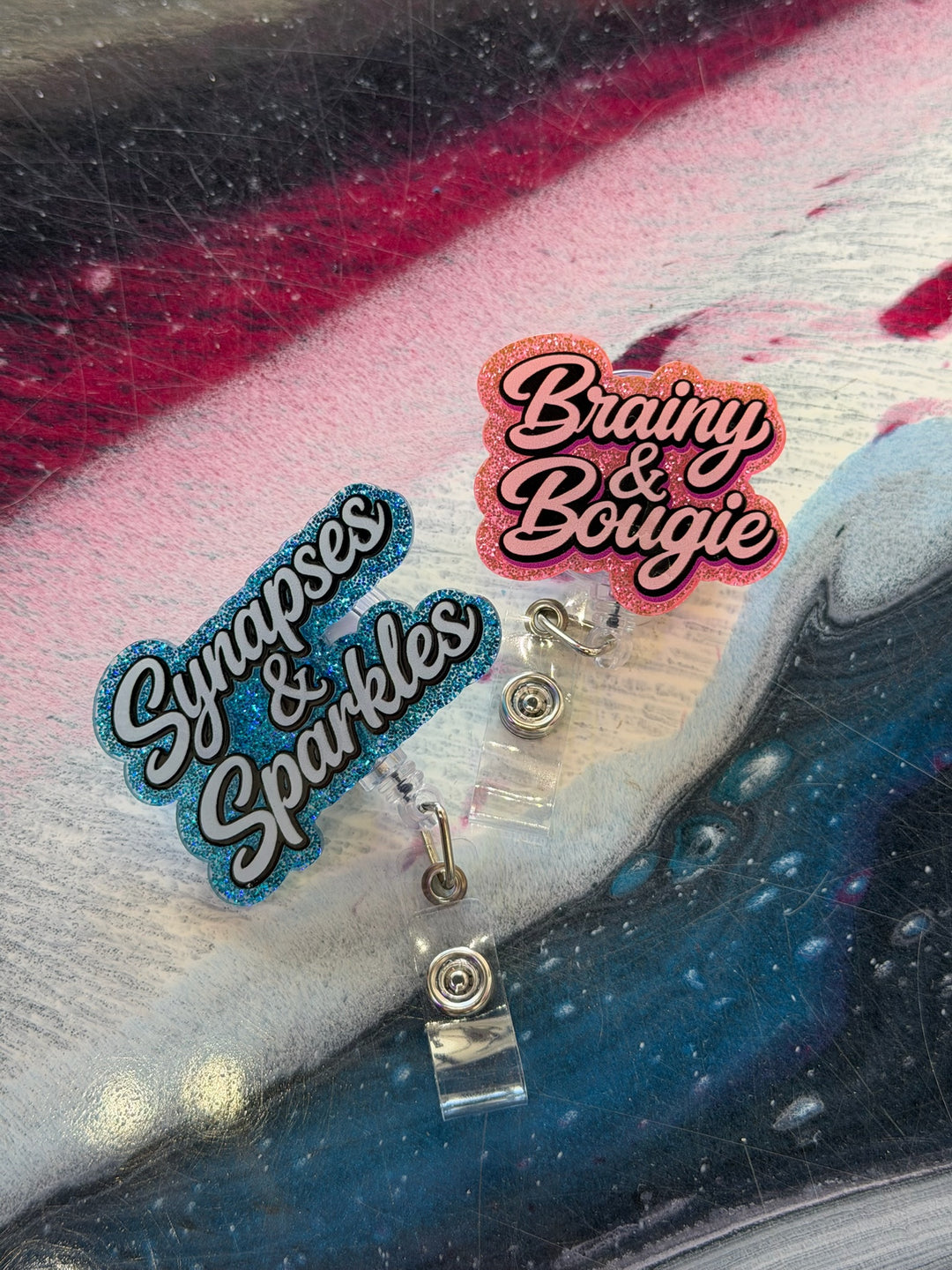 Two decorative badge reels on a textured surface with 'Synapses & Sparkles' and 'Brainy & Bougie' text.