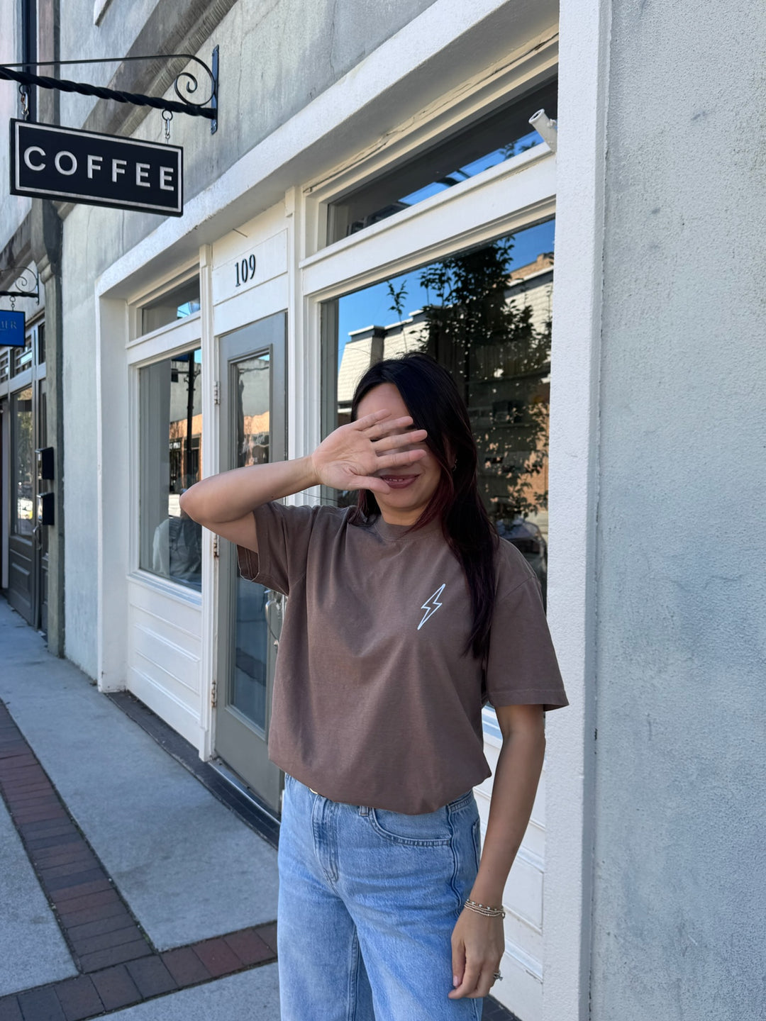 Person wearing The Incredible Tee Shirt - Brown Shirt with Motivational "YOU WILL DO INCREDIBLE THINGS. KEEP GOING." Motto and blue jeans standing in front of a coffee shop.