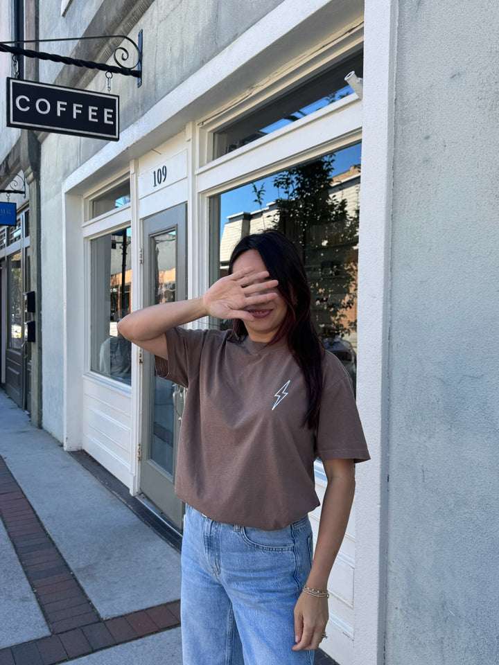 Person wearing The Incredible Tee Shirt - Brown Shirt with Motivational "YOU WILL DO INCREDIBLE THINGS. KEEP GOING." Motto and blue jeans standing in front of a coffee shop.