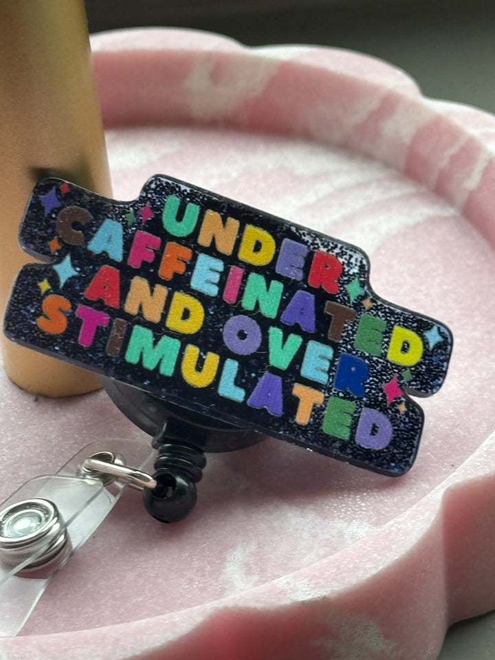 Retractable badge reel with colorful lettering that reads “Under Caffeinated and Over Stimulated” on a sparkly blue and black glitter background and displayed in a pink scallop tray.