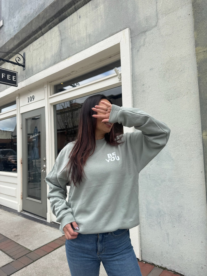 Woman wearing a sage green crewneck sweatshirt with “BE YOU.” printed on the front in bold white letters.