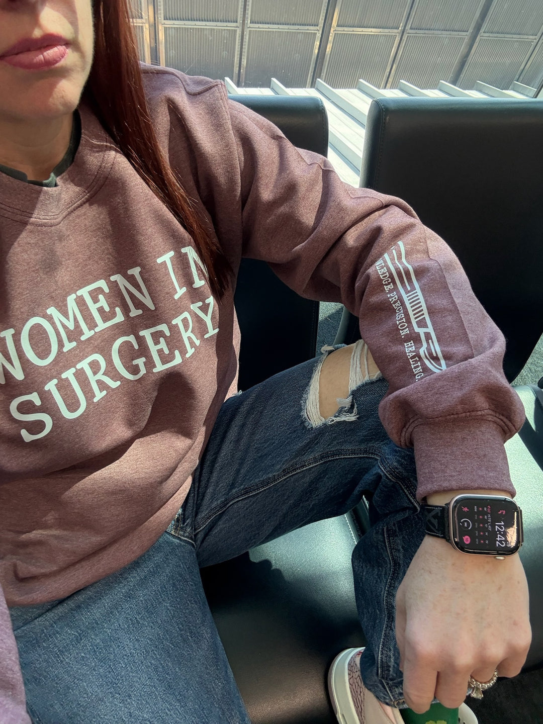 Dr. Betsy Grunch - Ladyspinedoc wearing a heather maroon crewneck sweatshirt with "WOMEN IN SURGERY" printed in bold white letters across the chest and showing off a white scalpel design with the words "KNOWLEDGE. PRECISION. HEALING." printed on the bottom left sleeve.