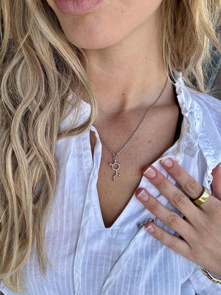 Close-up of a person wearing a white blouse with a silver Noradrenaline Molecule necklace and gold ring.