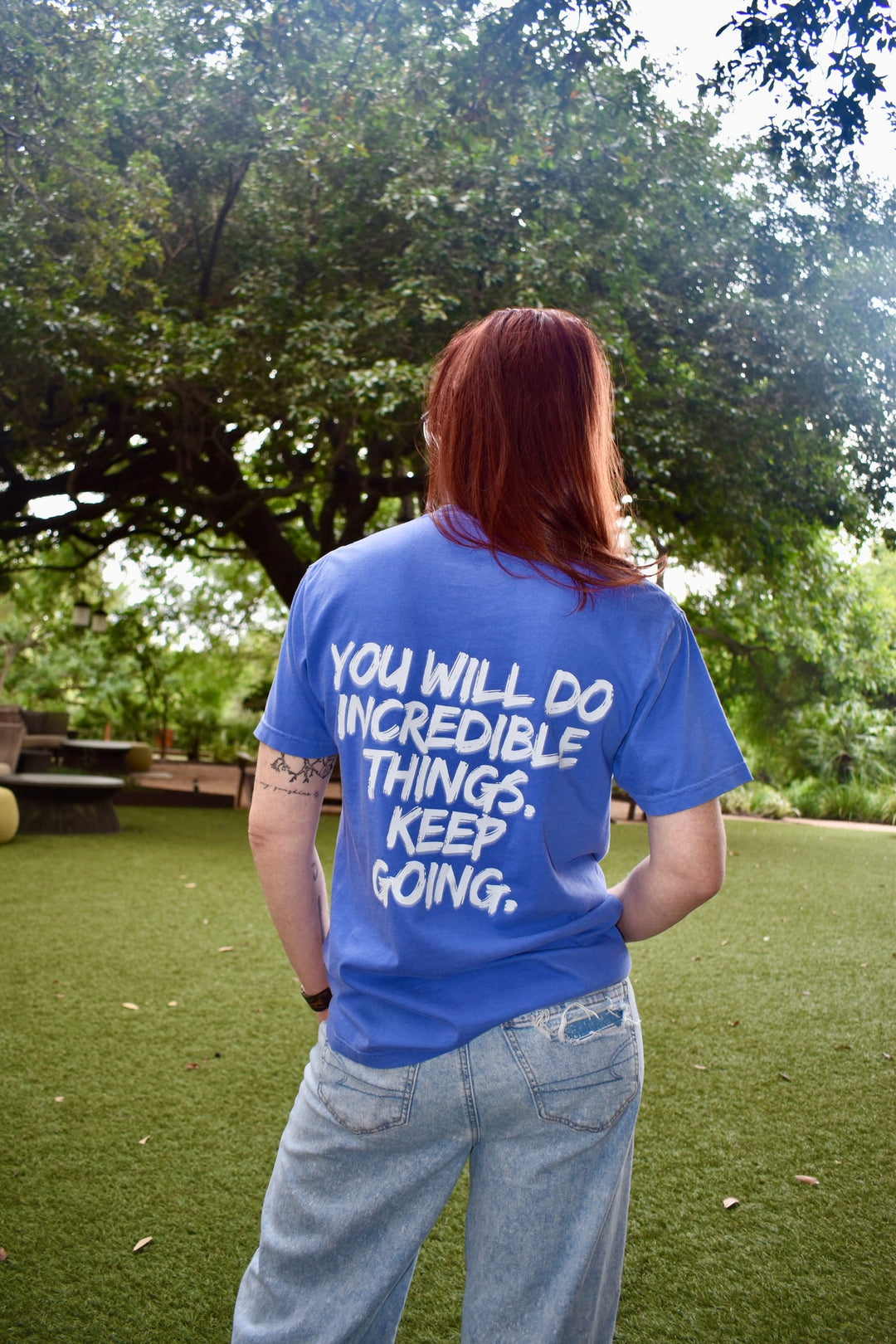 Full rear view of Ladyspinedoc in The Incredible Tee, message on back fully visible against a green outdoor backdrop.