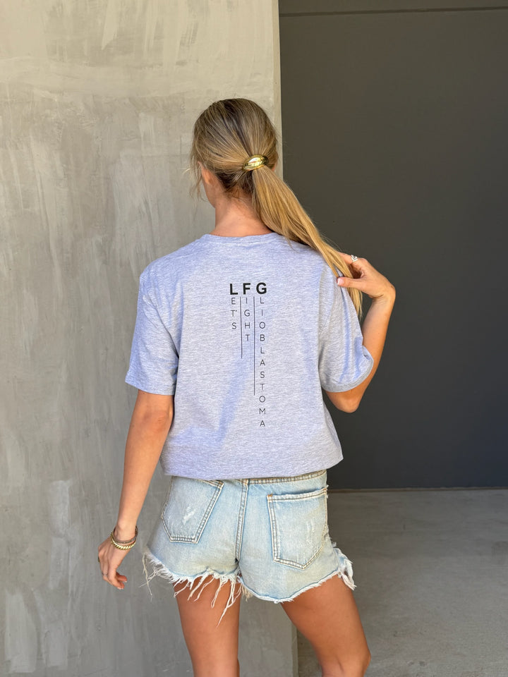 Person wearing a light gray t-shirt with LFG (Let's Fight Glioblastoma) text and denim shorts, standing against a plain wall.