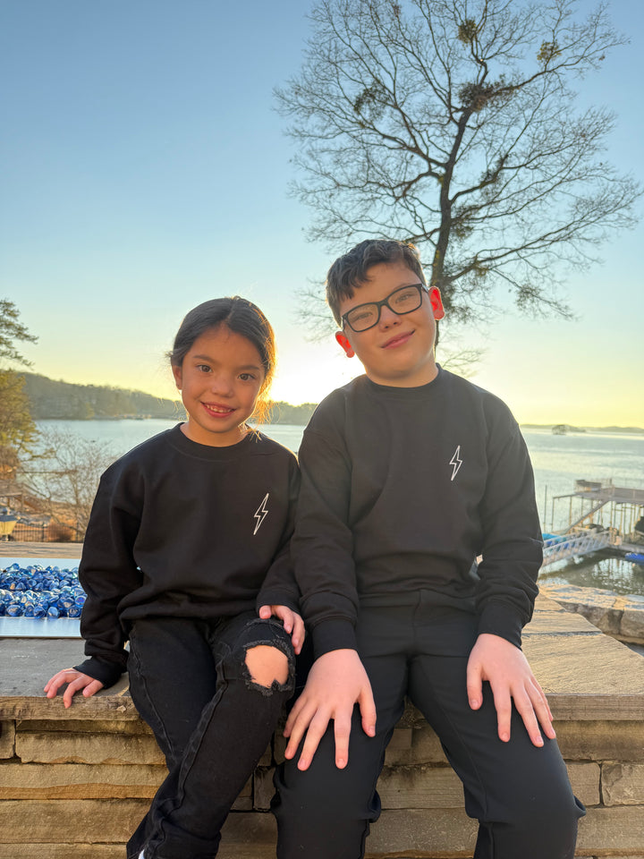 Youth models sitting and showing the front view of black Incredible Youth Crewneck Sweatshirt with white lightning bolt logo on chest.
