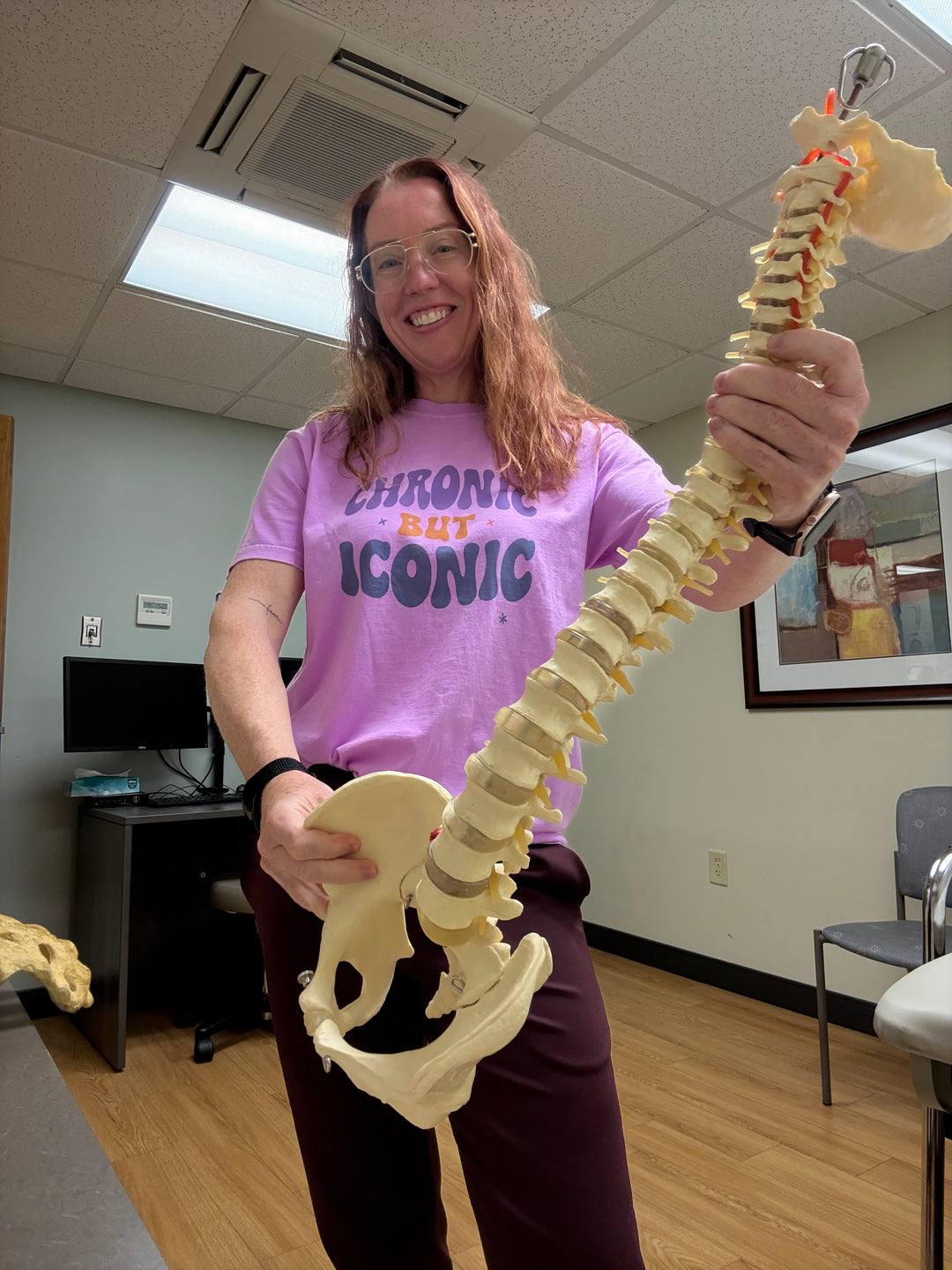 A smiling Dr. Betsy Grunch holding a model of a spine and wearing her Chronic But Iconic purple tee