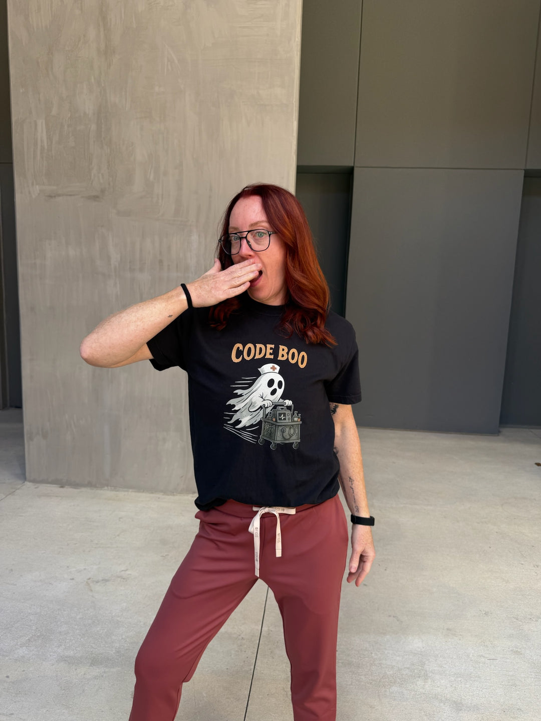 Ladyspinedoc wearing a black t-shirt with 'CODE BOO' and maroon pants standing against a plain wall.