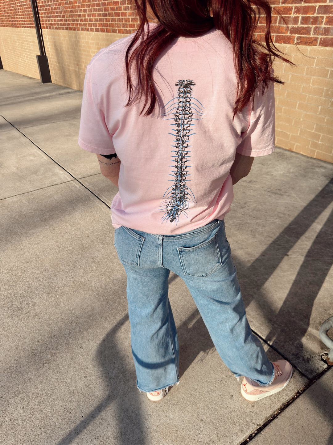 A person wearing a light pink short-sleeved tee shirt with a spine design on the back, standing with their back towards the camera.
