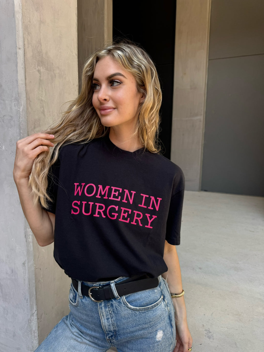 Woman wearing a black t-shirt with 'WOMEN IN SURGERY' in pink text, standing against a neutral wall.