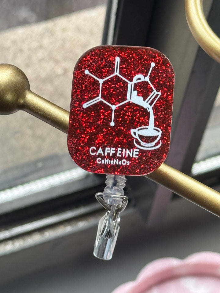 Ladyspinedoc Red Sparkly Enerjava Badge Reel - Retractable with Alligator Clip and Caffeine Molecule Design on a dark red glitter background, clipped to a gold stand.