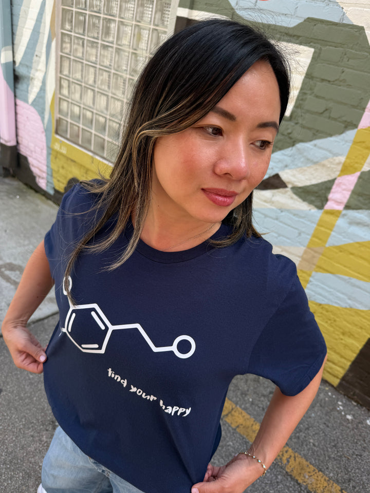 Woman wearing a navy blue t-shirt with a dopamine molecule design and text, standing in front of a colorful wall.