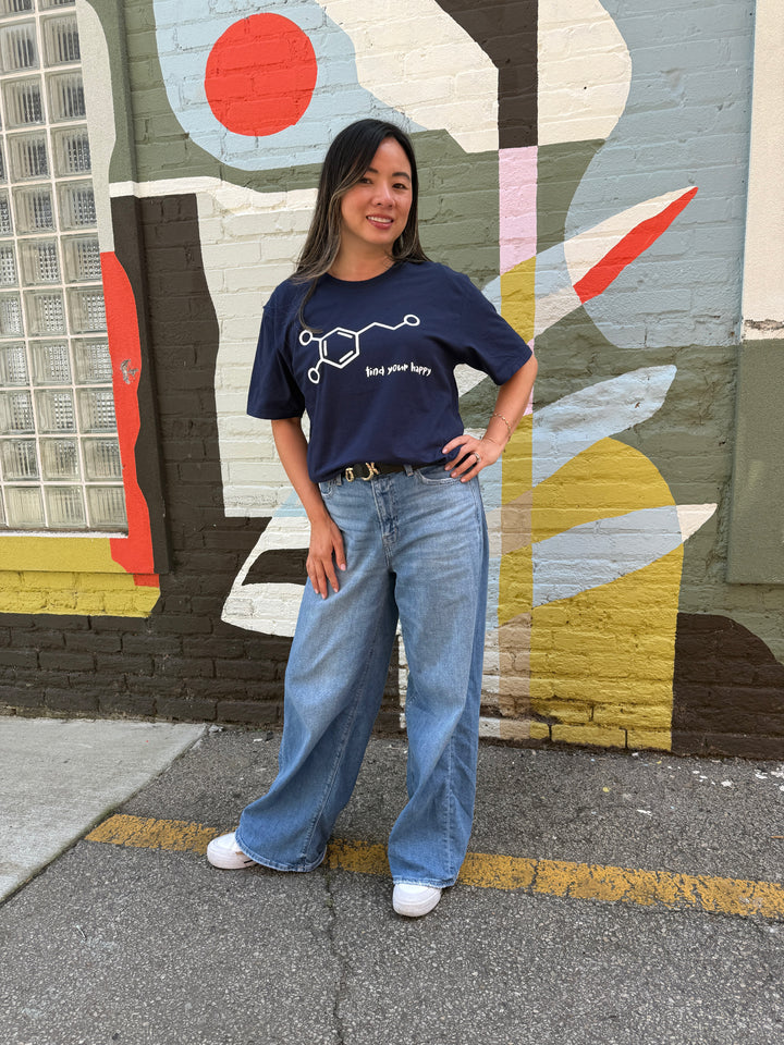 Person wearing a blue dopamine molecule t-shirt and jeans standing in front of a colorful mural.