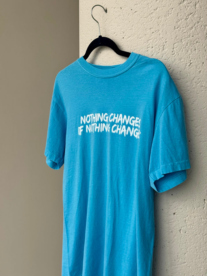 “Nothing Changes If Nothing Changes” Blue Tee Shirt hanging on hanger on a cement wall. 