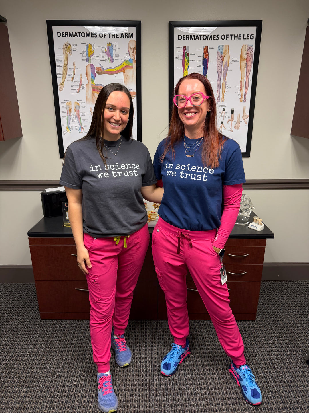 two ladies standing in a doctor's office in navy and gray shirts that say "in science we trust" on them