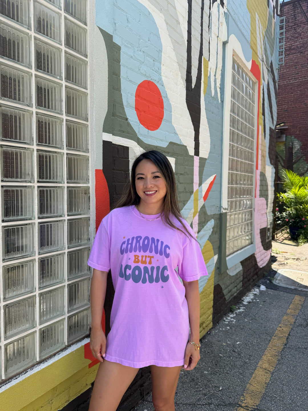 Woman wearing a purple Chronic But Iconic t-shirt with text standing in front of a colorful mural.