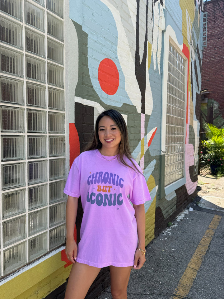 Woman wearing a purple Chronic But Iconic t-shirt with text standing in front of a colorful mural.