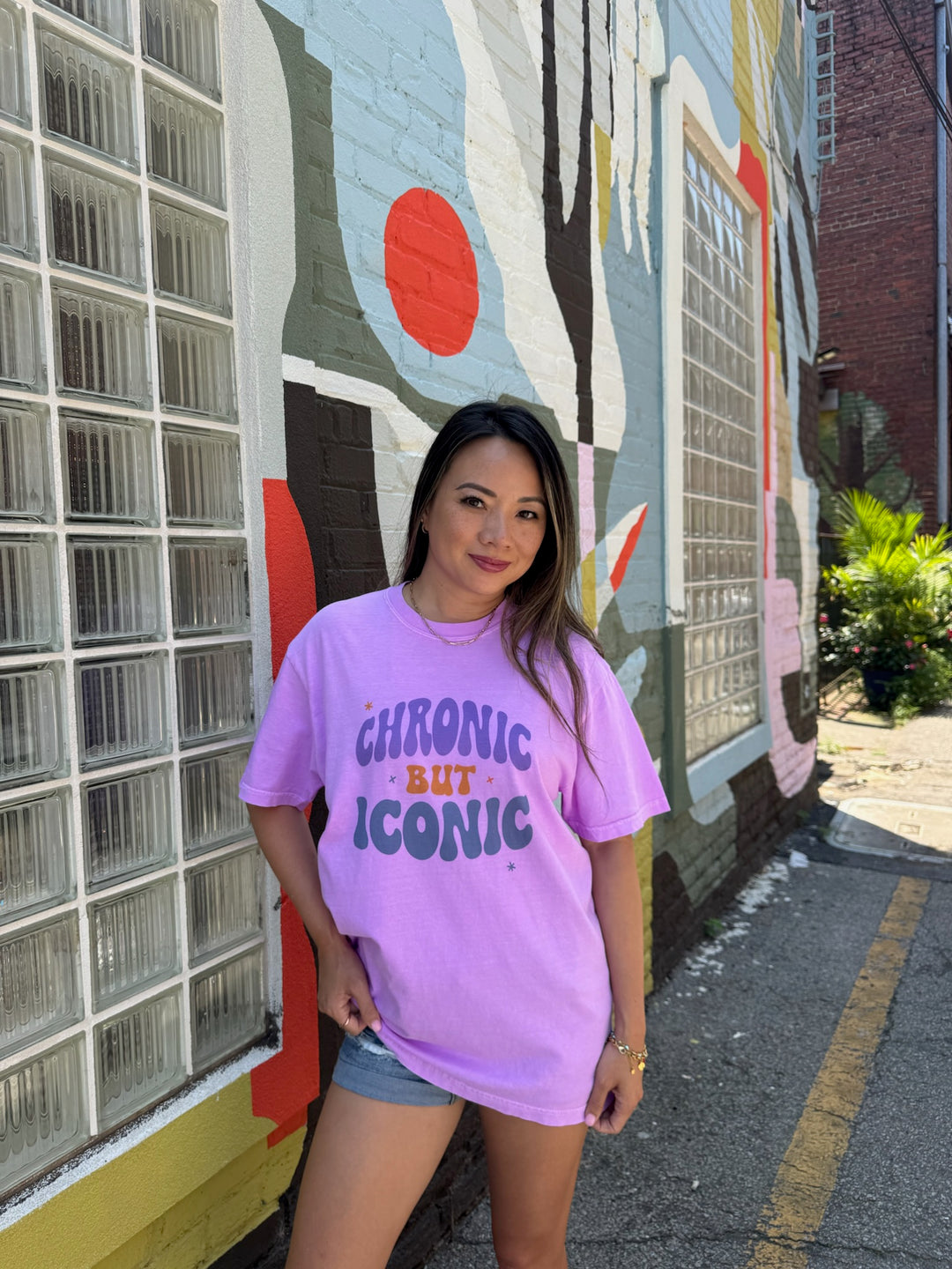 Woman wearing a purple t-shirt with 'Chronic But Iconic' text in front of a colorful mural.