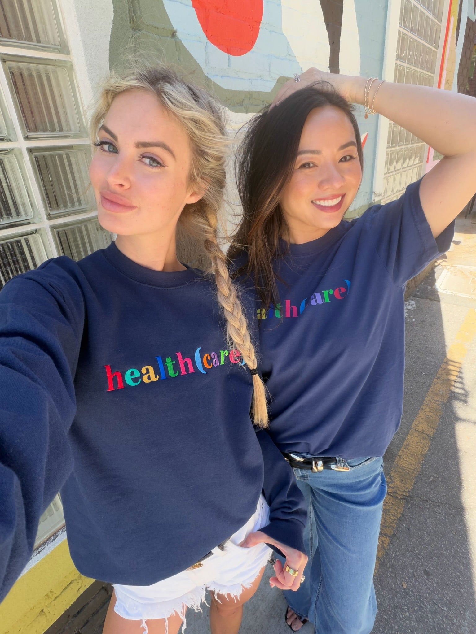 Two women wearing navy blue sweatshirts with colorful "health(care)", standing on a street.