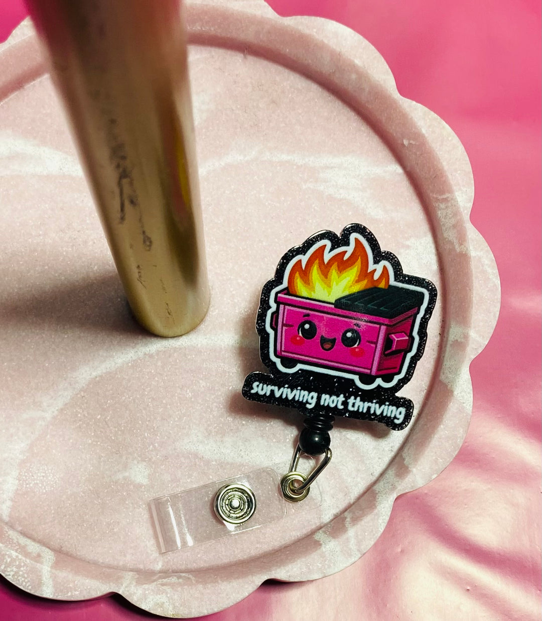 Retractable badge holder with a cartoon dumpster on fire design and 'surviving not thriving' text, in a pink tray