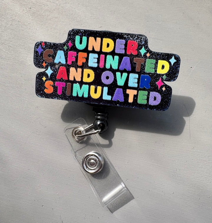 Retractable badge reel with bold, colorful lettering that reads “Under Caffeinated and Over Stimulated” on a sparkly blue and black glitter background. 