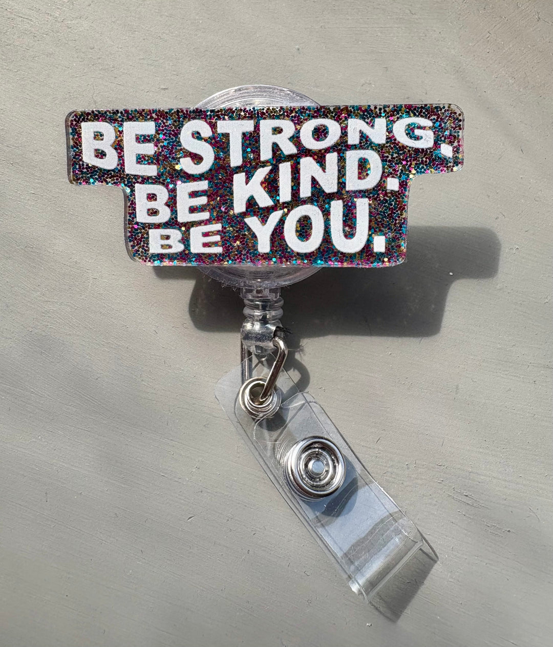 Retractable badge reel with bright multicolor glitter background and bold white text that reads "BE STRONG. BE KIND. BE YOU." with a clear name ID clip attached.