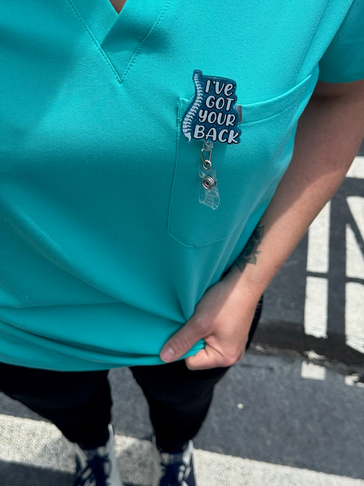 Blue Glitter I'VE GOT YOUR BACK Spine Badge Reel - Retractable with Alligator Clip clipped on front pocket of aqua scrubs