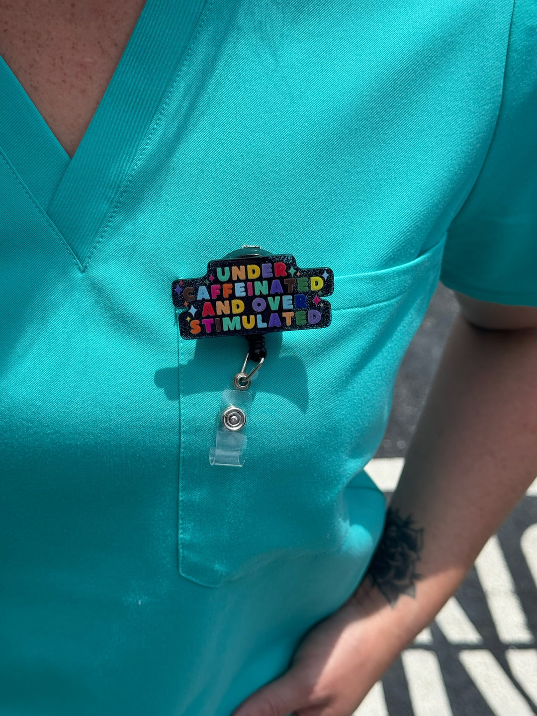 Under Caffeinated and Over Stimulated Badge Reel clipped on front pocket of scrub shirt
