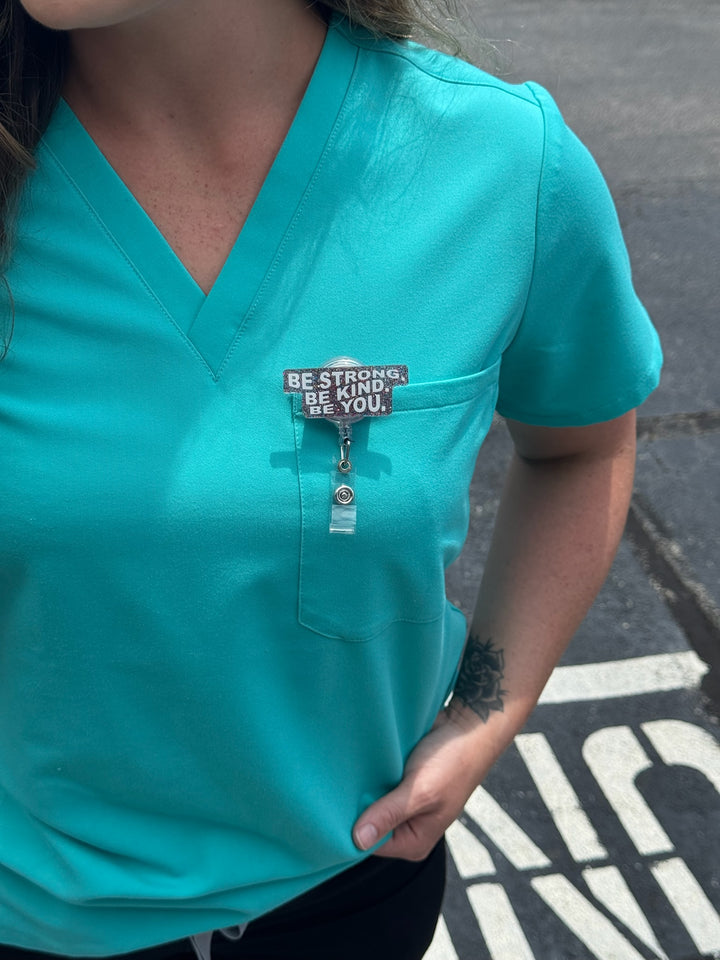 Be Strong. Be Kind. Be You. Glitter Badge Reel - Retractable with Alligator Clip shown clipped on front pocket of scrubs