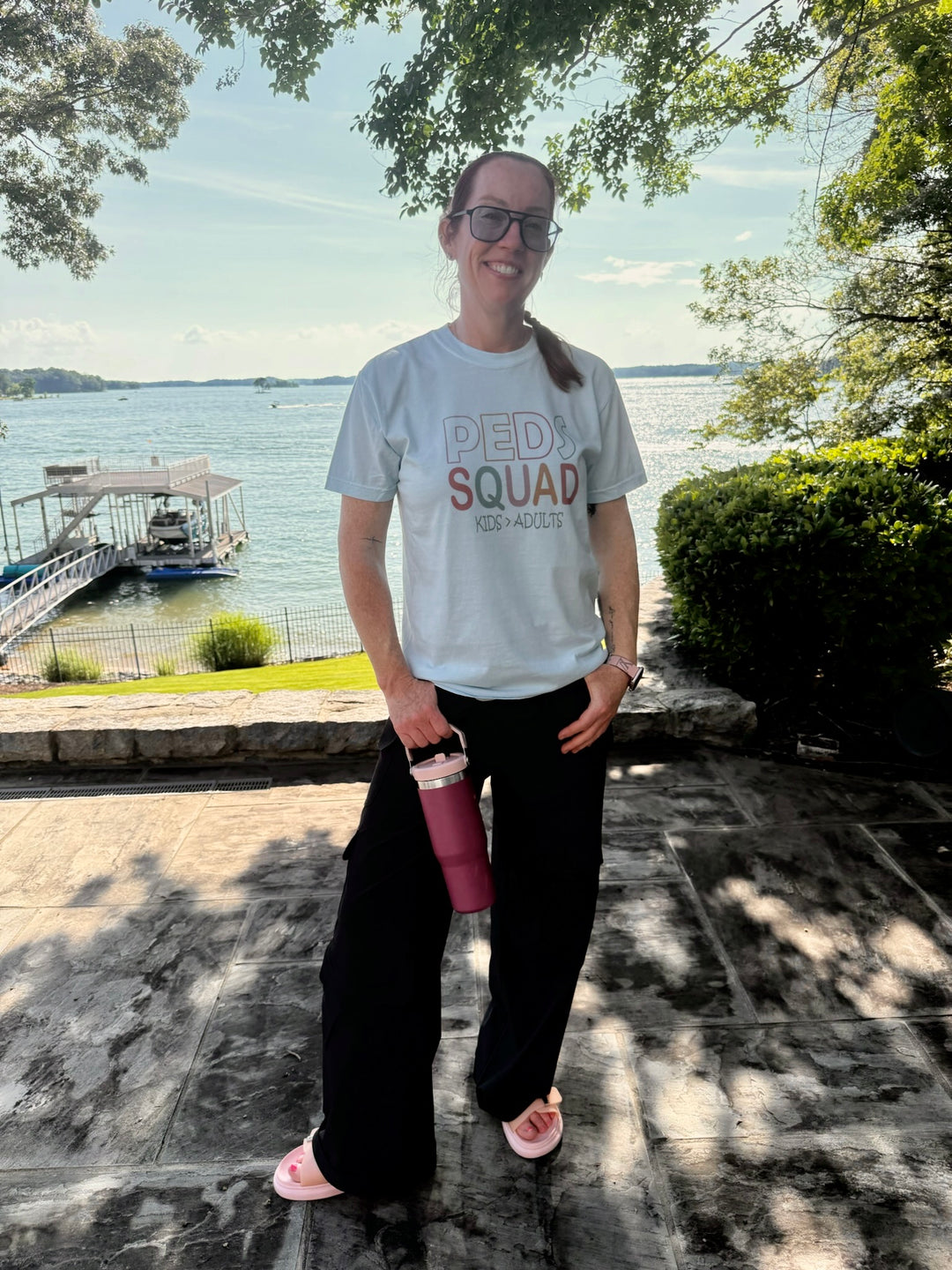 Dr. Betsy Grunch stands on a sunlit patio near a lake, wearing a pastel blue “PEDS SQUAD – KIDS > ADULTS” t-shirt. She smiles at the camera, holding a pink water bottle, with a boat dock and trees in the background.